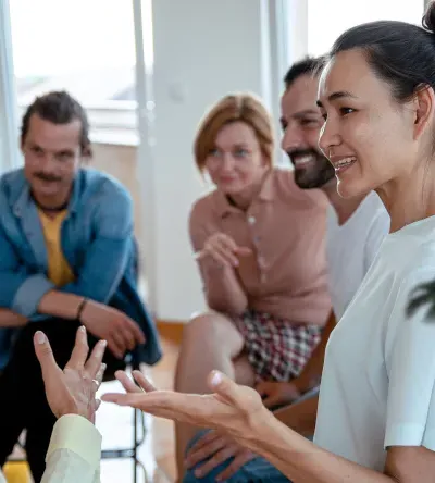 A diverse group of people is seated in a circle in a group therapy or support session. A woman in white is speaking, and others, including a man and a woman in a red shirt, listen and engage with her. The room has bright windows and a warm atmosphere.